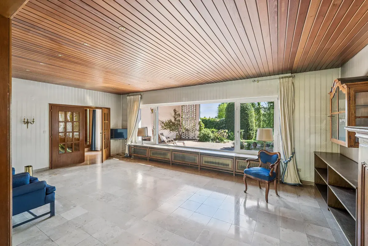 Bright living room with wood ceiling, marble floor, and large window overlooking a garden. Blue chair and sofa add color.