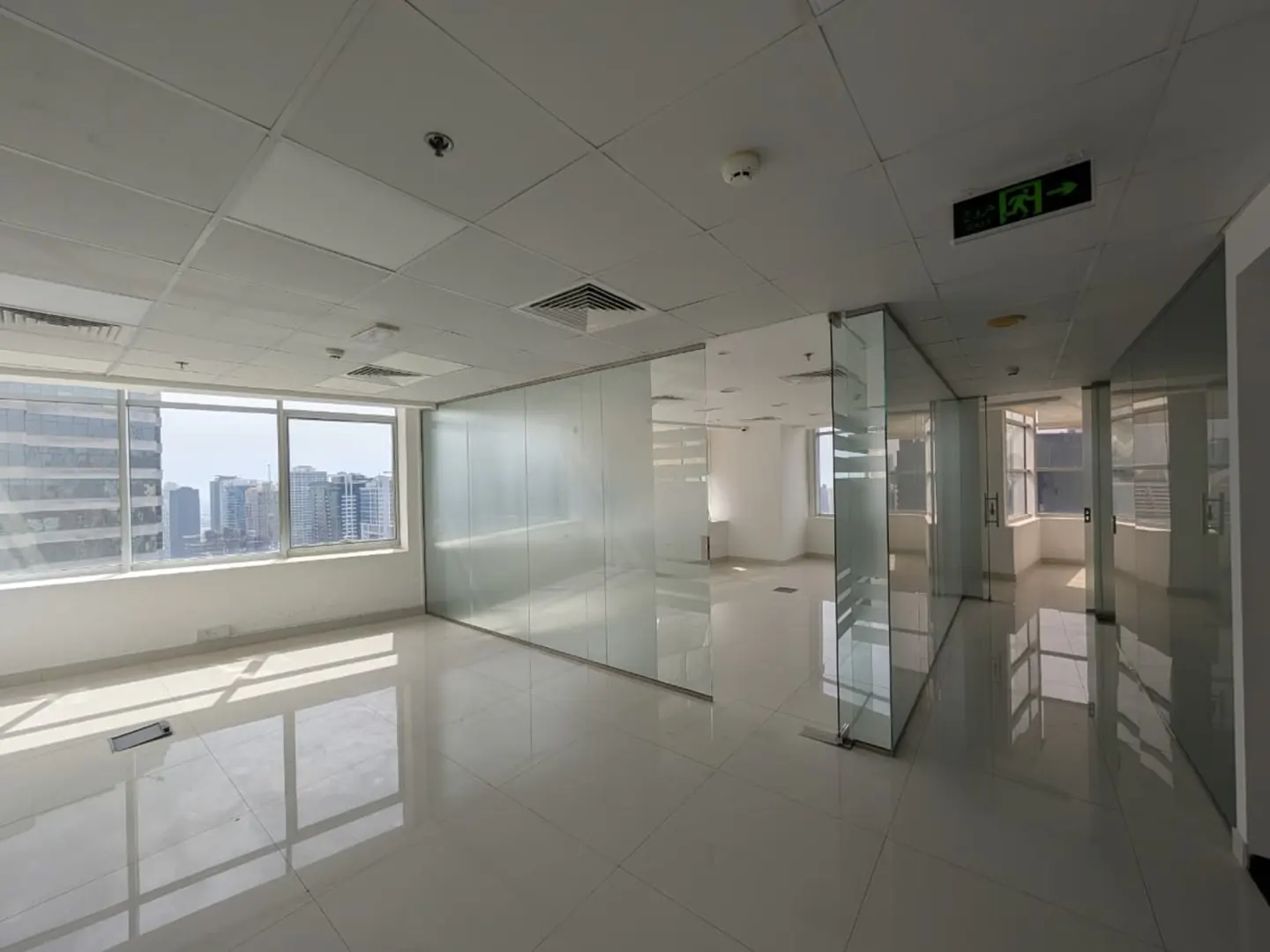 Bright, empty office space with white tile floors, glass walls, and large windows overlooking a city skyline. An exit sign is visible on the ceiling.