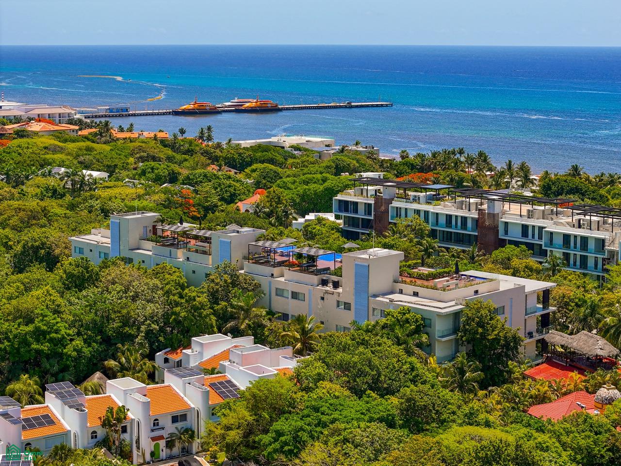 Aerial view of modern white apartment buildings nestled in lush green trees near a blue ocean with a pier and ferry boats. [AI-generated text]