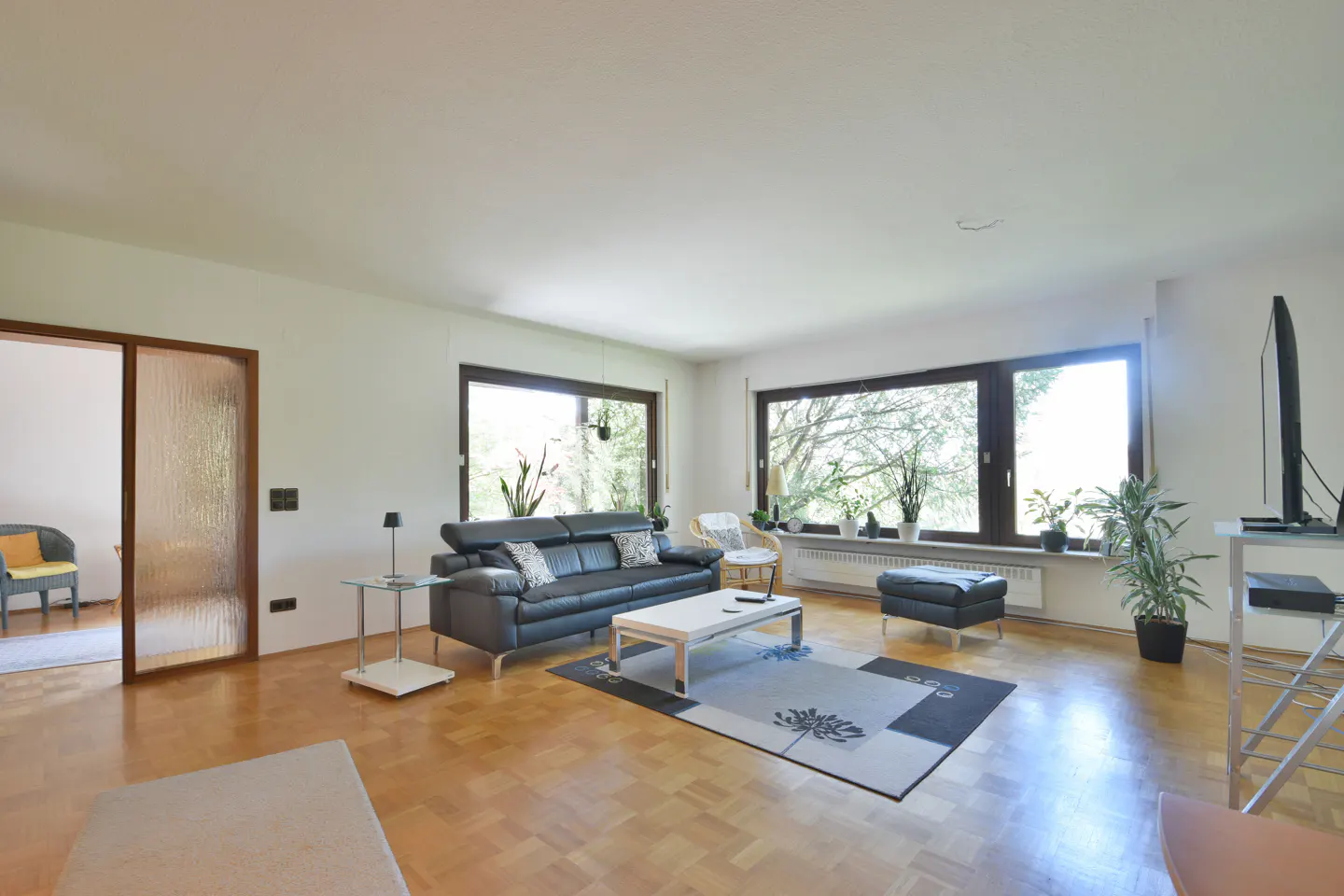 Bright living room with parquet floors, black leather sofa, white coffee table on a floral rug, and large windows.