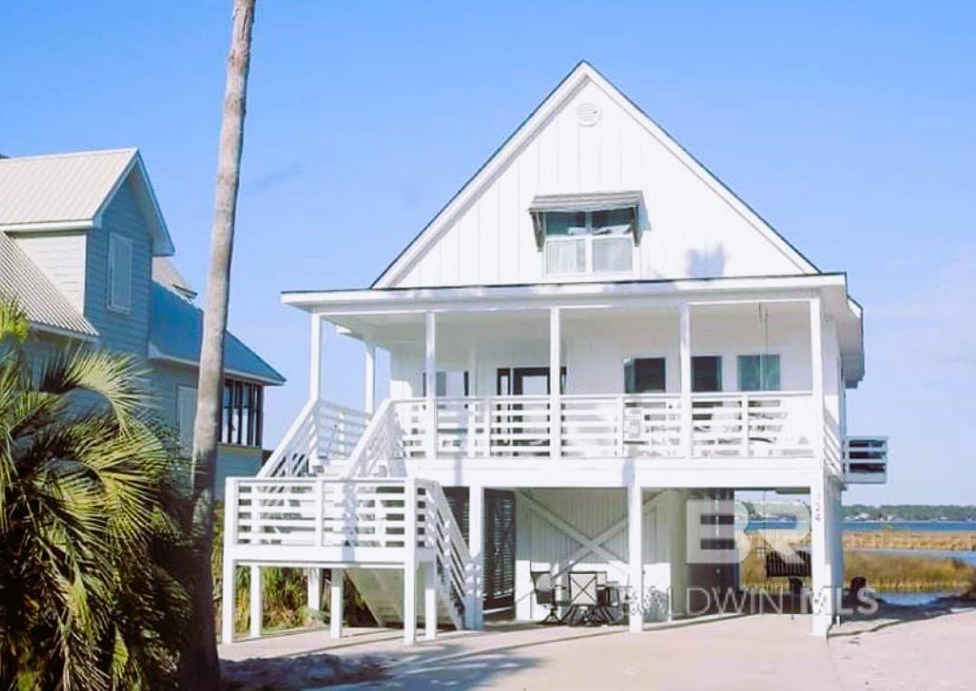 A white, two-story beach house on stilts with a porch and stairs, under a clear blue sky.