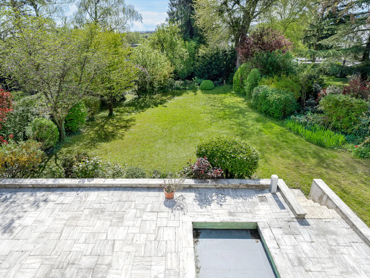 View from a stone patio overlooking a green lawn surrounded by trees and bushes. A potted plant sits on the patio.