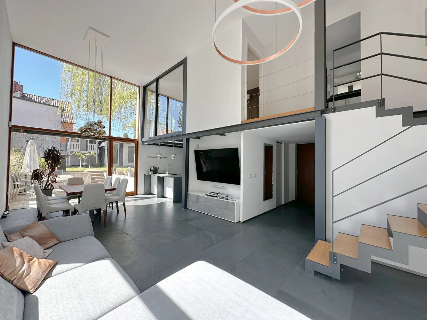 Bright, modern living room with gray sofa, dining area, TV, and open staircase. Large windows offer outdoor views. Circular light fixture overhead.