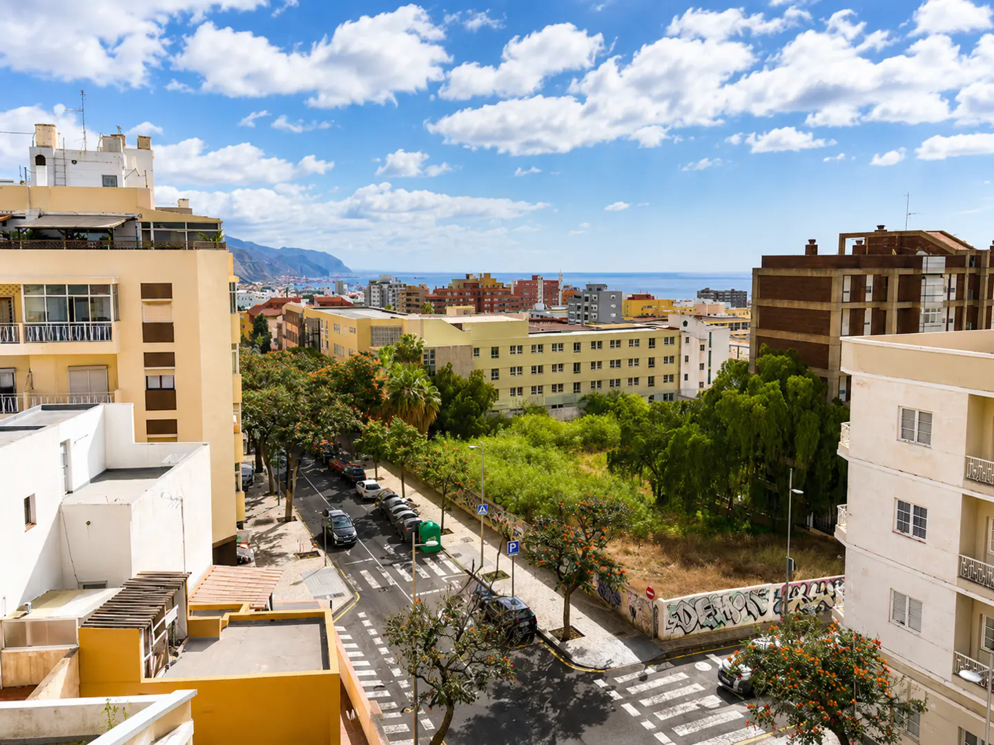 Cityscape view with buildings, trees, and a street with cars. Blue sky with white clouds in the background.