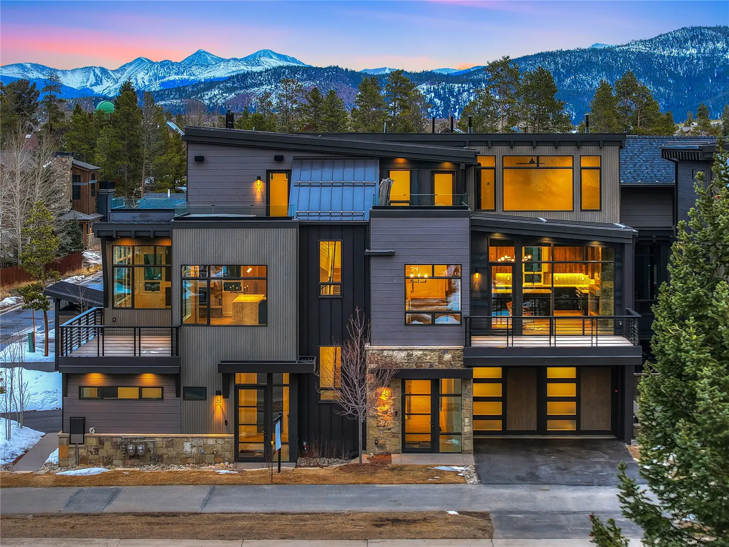 Modern gray house with large windows, set against a backdrop of snow-capped mountains and a twilight sky.