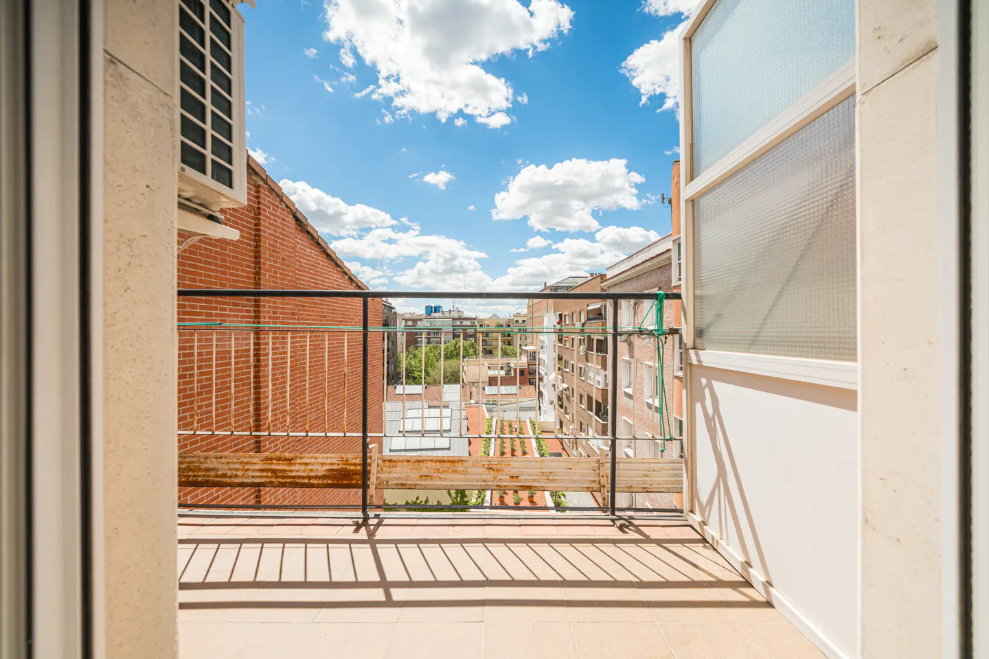 View from a balcony with black railings, showing brick buildings and a blue sky with white clouds. An open window is on the right.