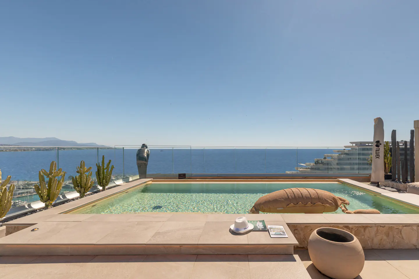 Rooftop pool with ocean view. A statue looks out over the water. A hat and book sit poolside. A tan float is in the water.