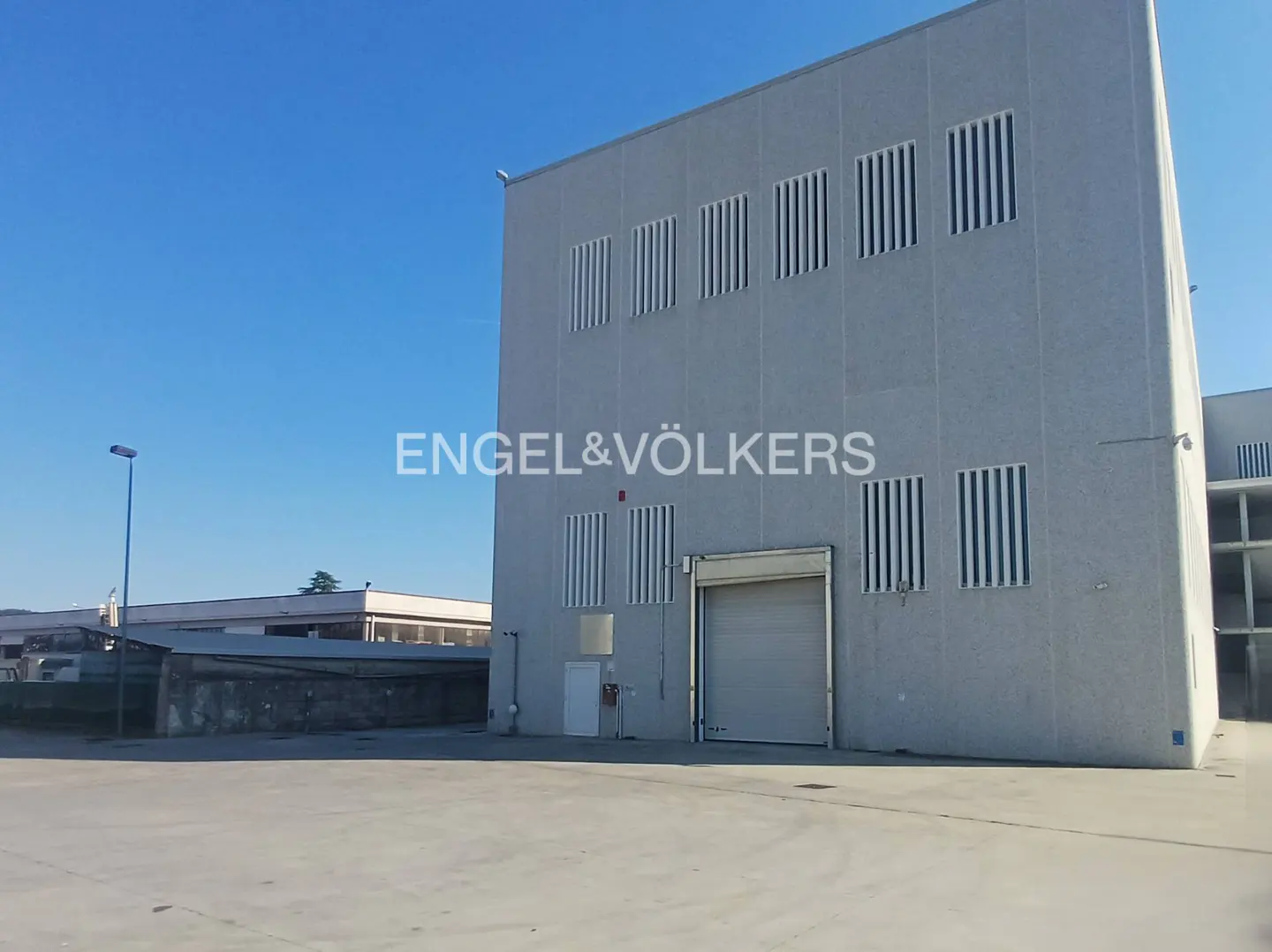 Tall, light gray industrial building with a large roll-up door and vertical windows under a clear blue sky.