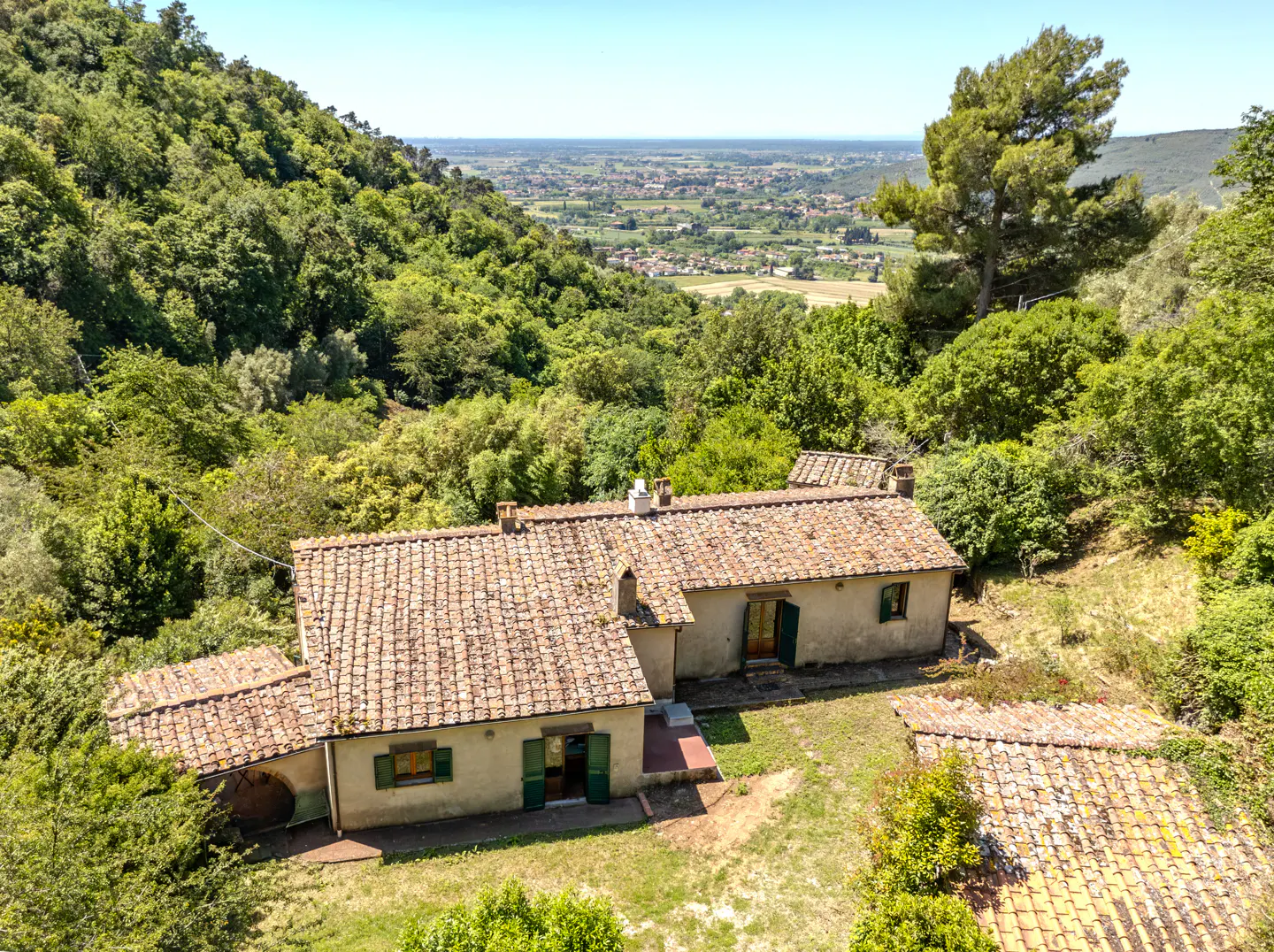 Aerial view of a tan house with a terracotta tile roof, green shutters, surrounded by lush green trees and a valley in the background.