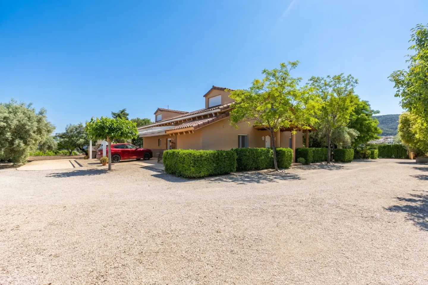 Tan house with a red tile roof, green bushes, and a red car parked in the driveway.