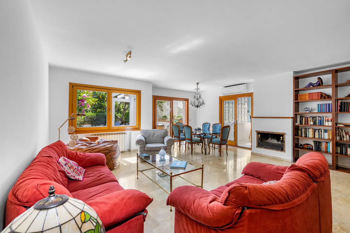 Bright living room with two red sofas, a glass coffee table, a dining area, and a large wooden bookshelf with a fireplace.