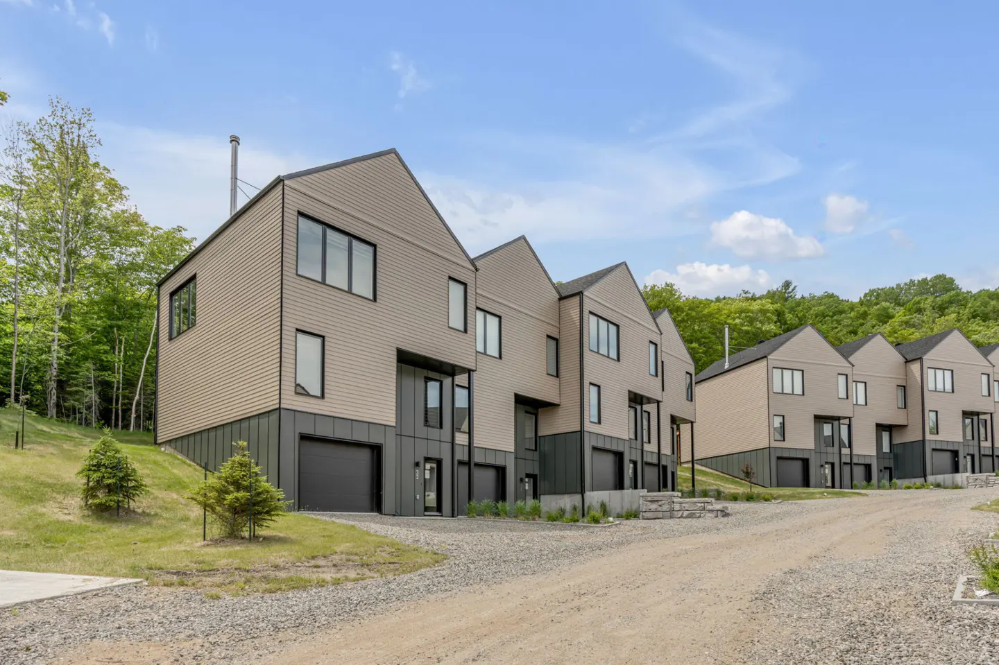 Row of modern townhouses with beige siding, black trim, and garage doors on a gravel driveway near a wooded area.
