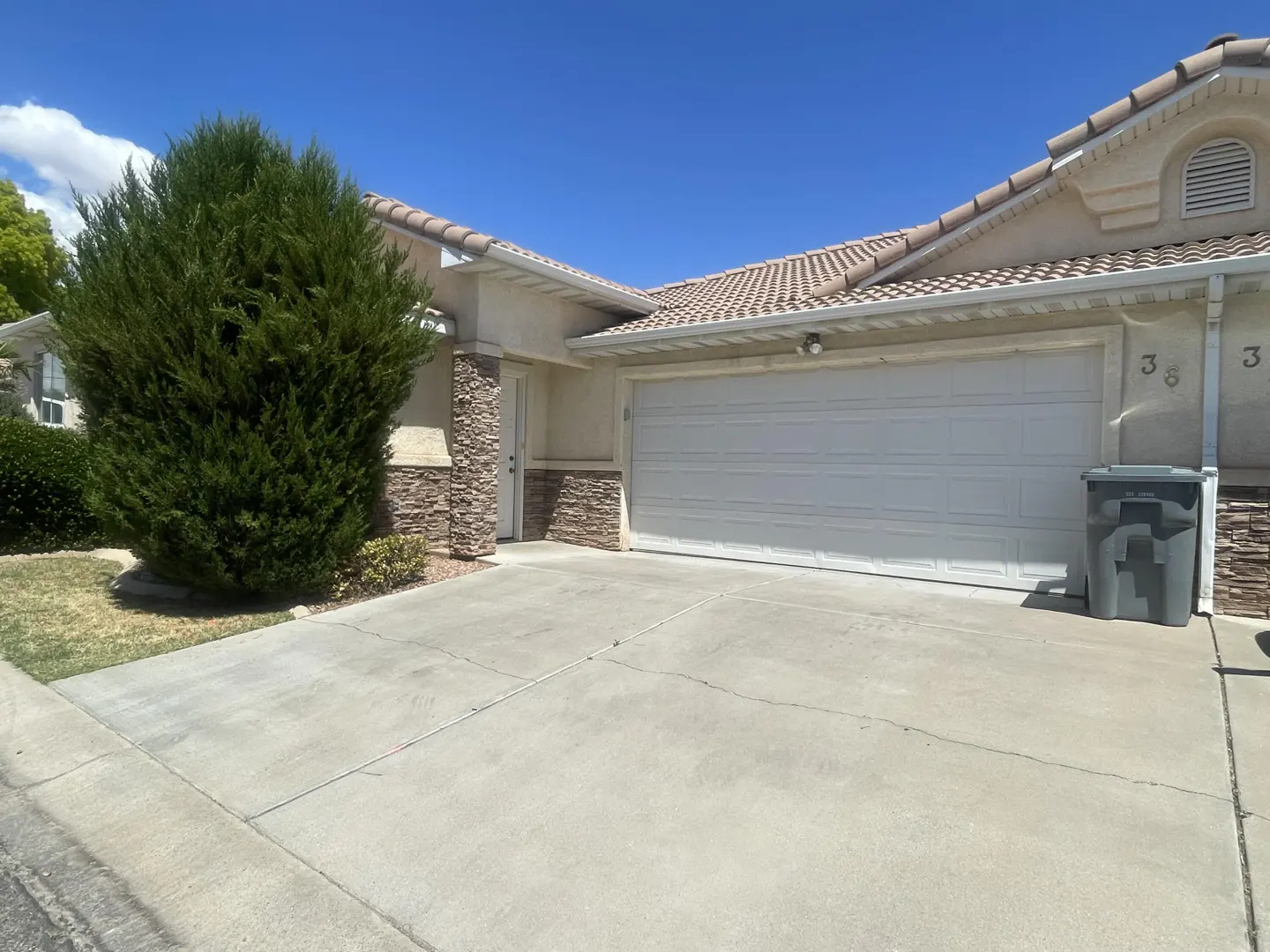 Beige single-story house with a white garage door, stone accents, and a green bush. A gray trash can sits beside the house. Blue sky.
