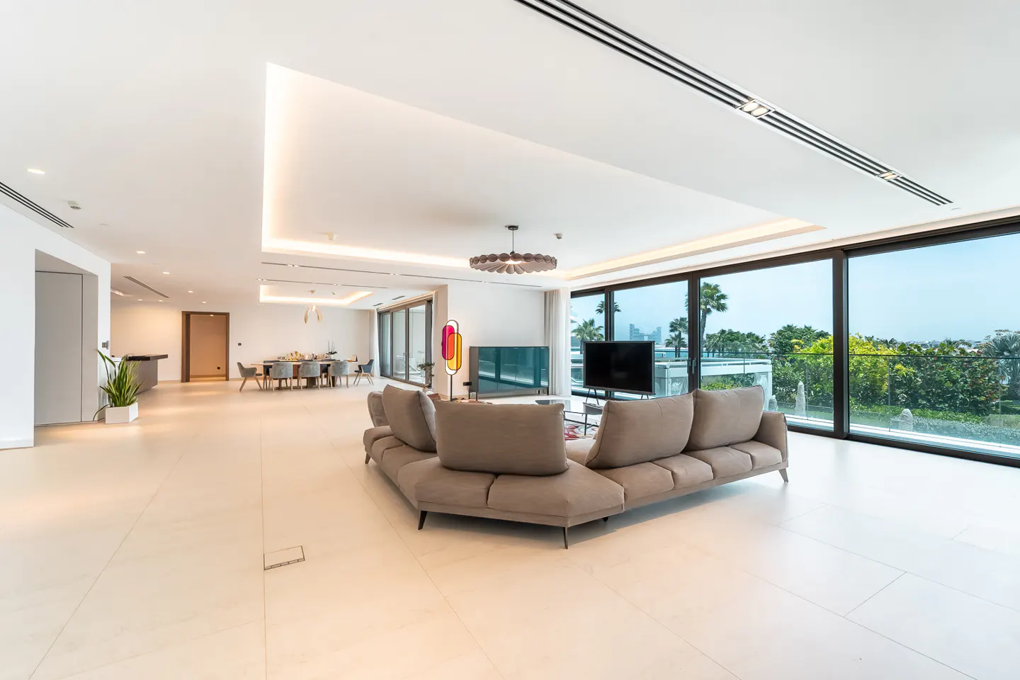 Bright, modern living room with a large gray sofa, TV, and floor-to-ceiling windows showing a green outdoor view.
