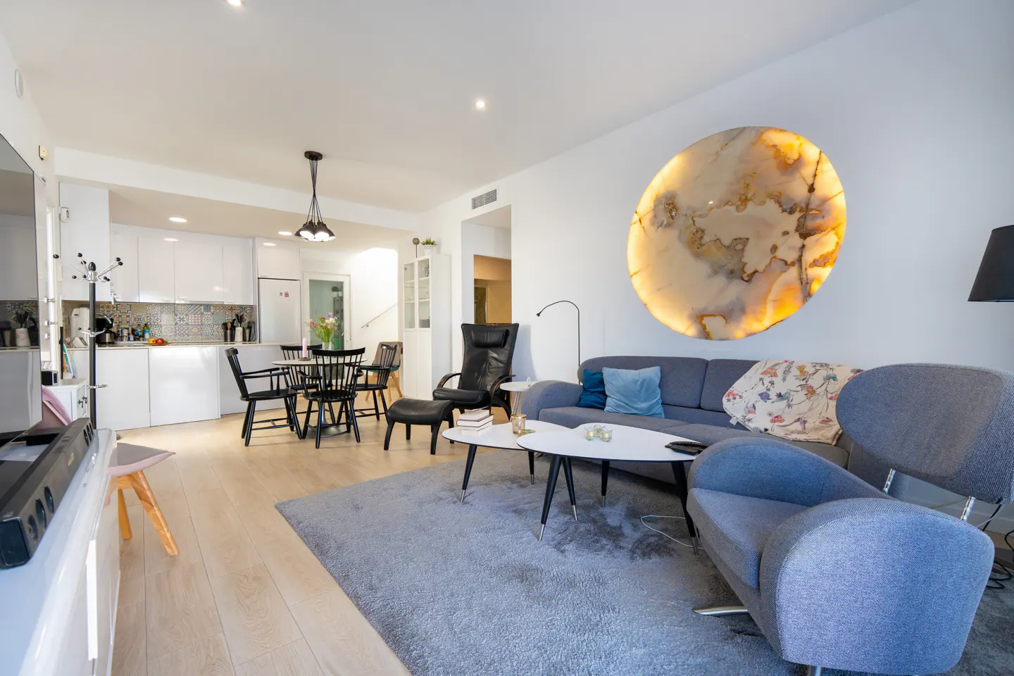 Modern living room with a grey sofa, blue armchair, and a large, circular backlit stone art piece on a white wall.