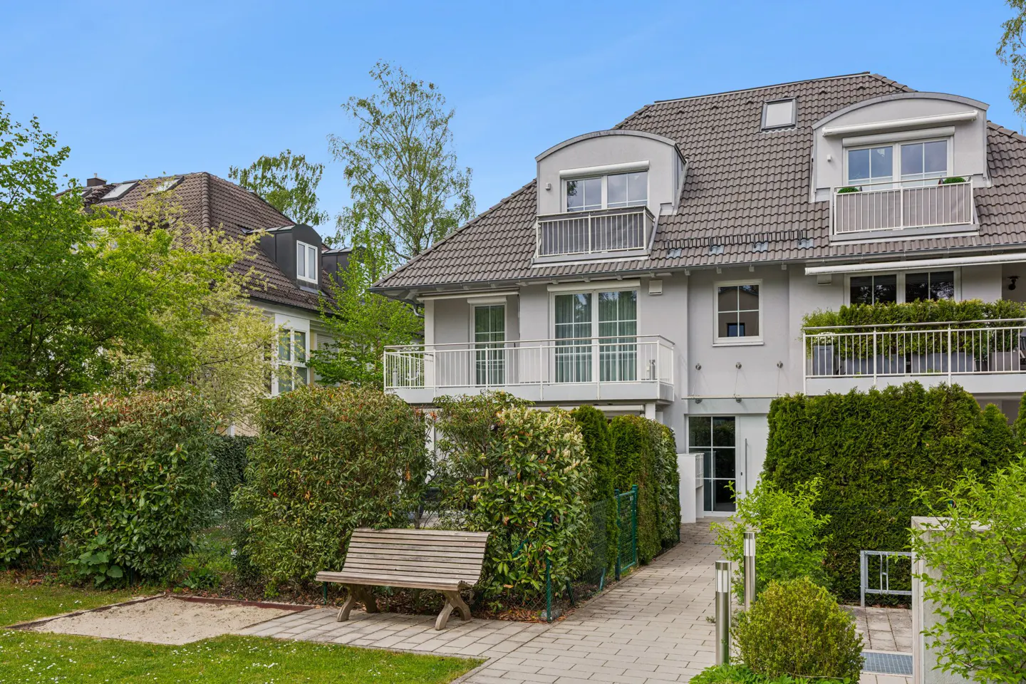 Exterior view of a gray, multi-story townhouse with balconies, a tiled roof, and a garden with a bench.