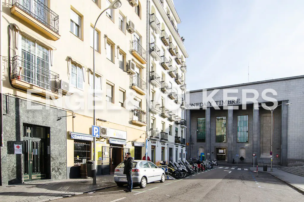 Street view of a light-colored building with balconies, a parked car, and a row of scooters. A man stands near the car.