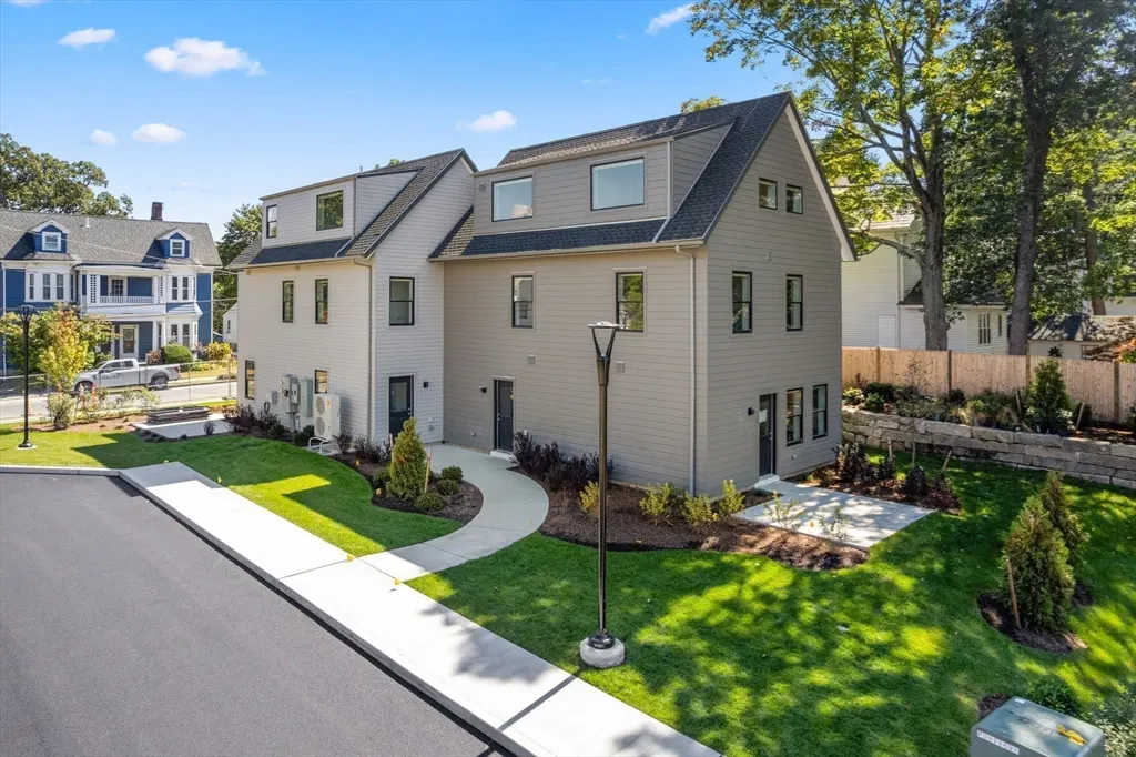 Exterior view of a modern, two-story home with gray siding, black trim, and a curved walkway leading to the entrance. Green lawn and trees surround the property.
