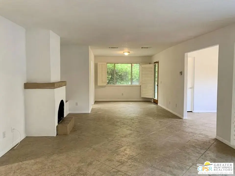 Bright, empty living room with white walls, tile floor, and a white fireplace. A window with white shutters looks out onto green trees.