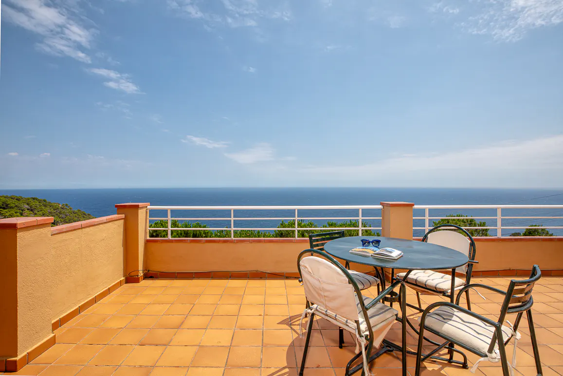 A sunny terrace with a round table and chairs overlooking a vast blue ocean under a clear sky.