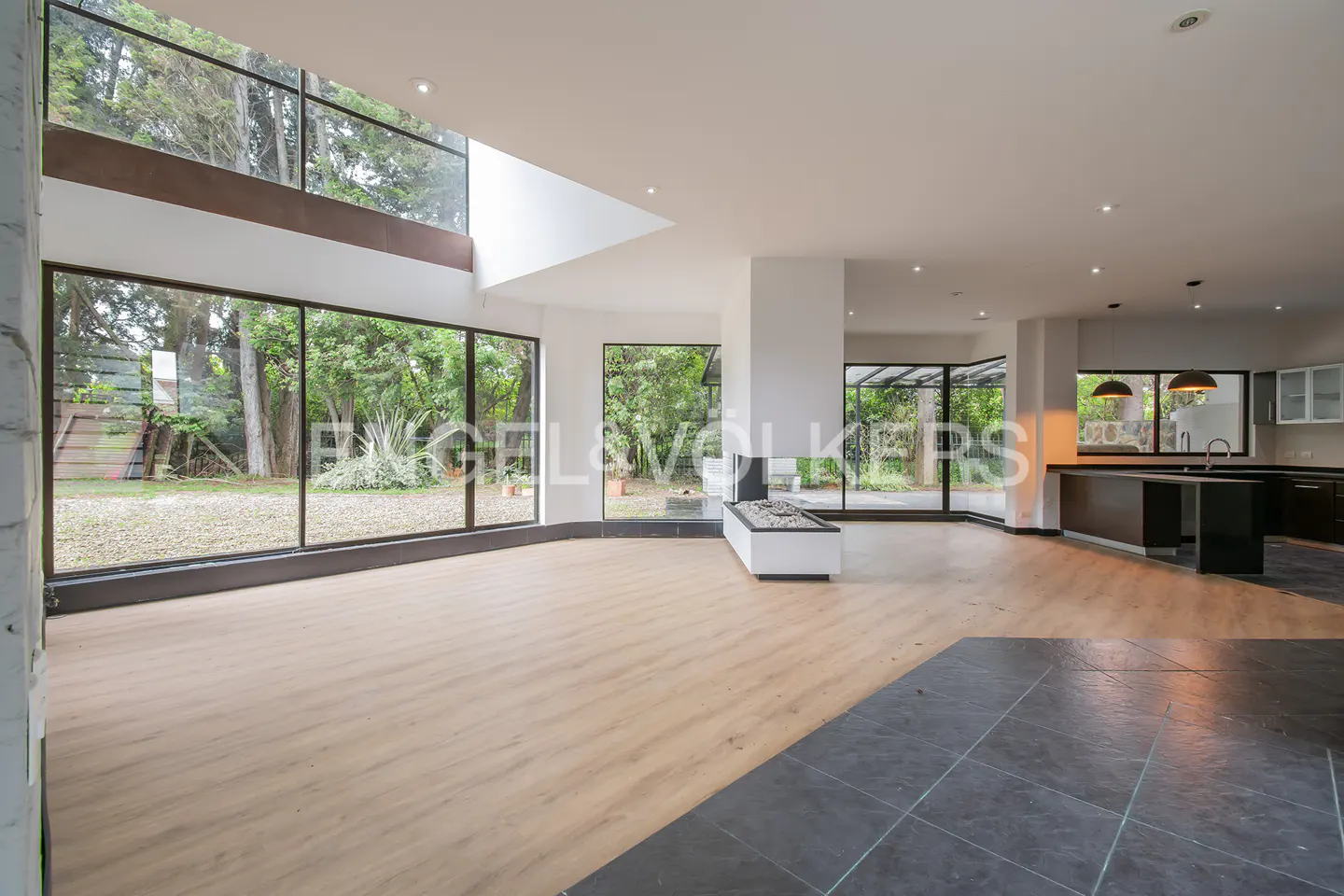 Bright, open-concept living space with wood floors, black tile, and floor-to-ceiling windows overlooking a lush green yard. Modern fireplace and kitchen island.