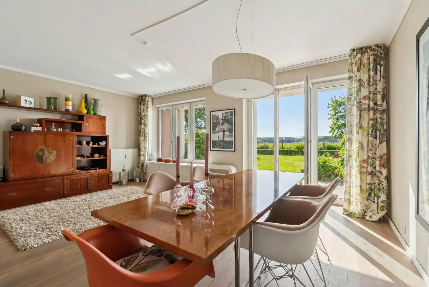 Bright dining room with a wood table, modern chairs, and a large cabinet. Windows overlook a green lawn.