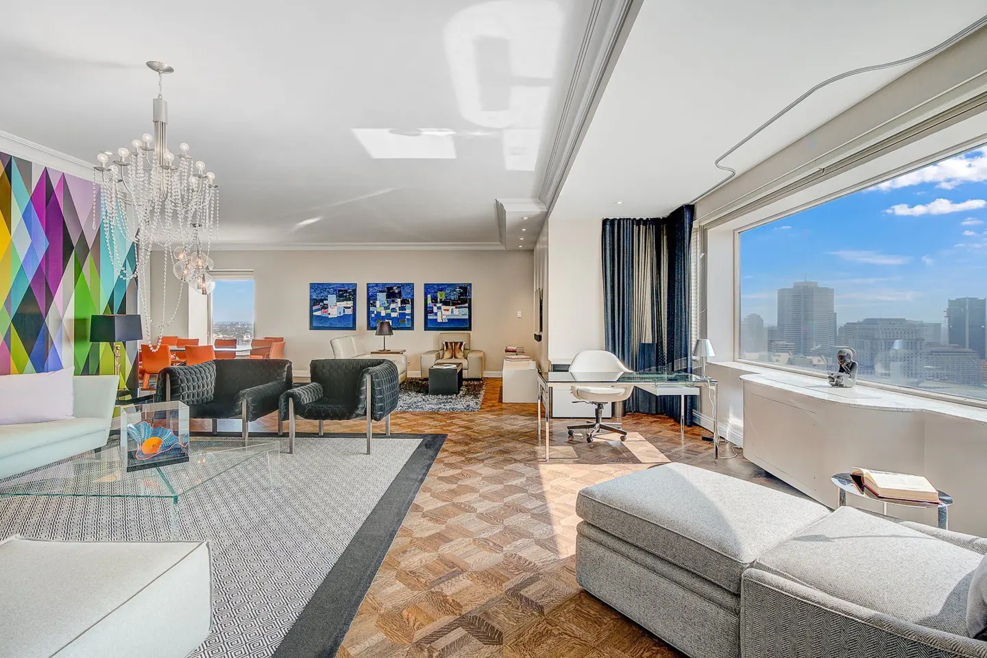 Bright, modern living room with a large window overlooking a city skyline. Features include a chandelier, colorful wall art, and stylish furniture.