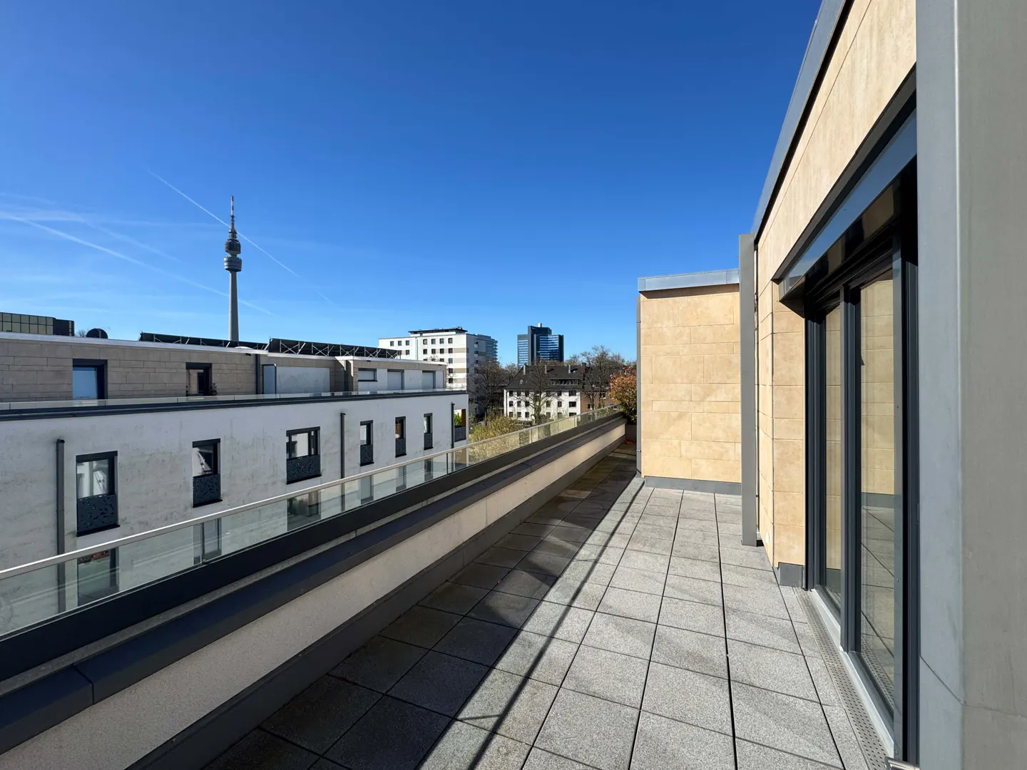 View from a balcony with gray tiles and glass railing, overlooking city buildings under a clear blue sky. A tall tower is visible in the distance.