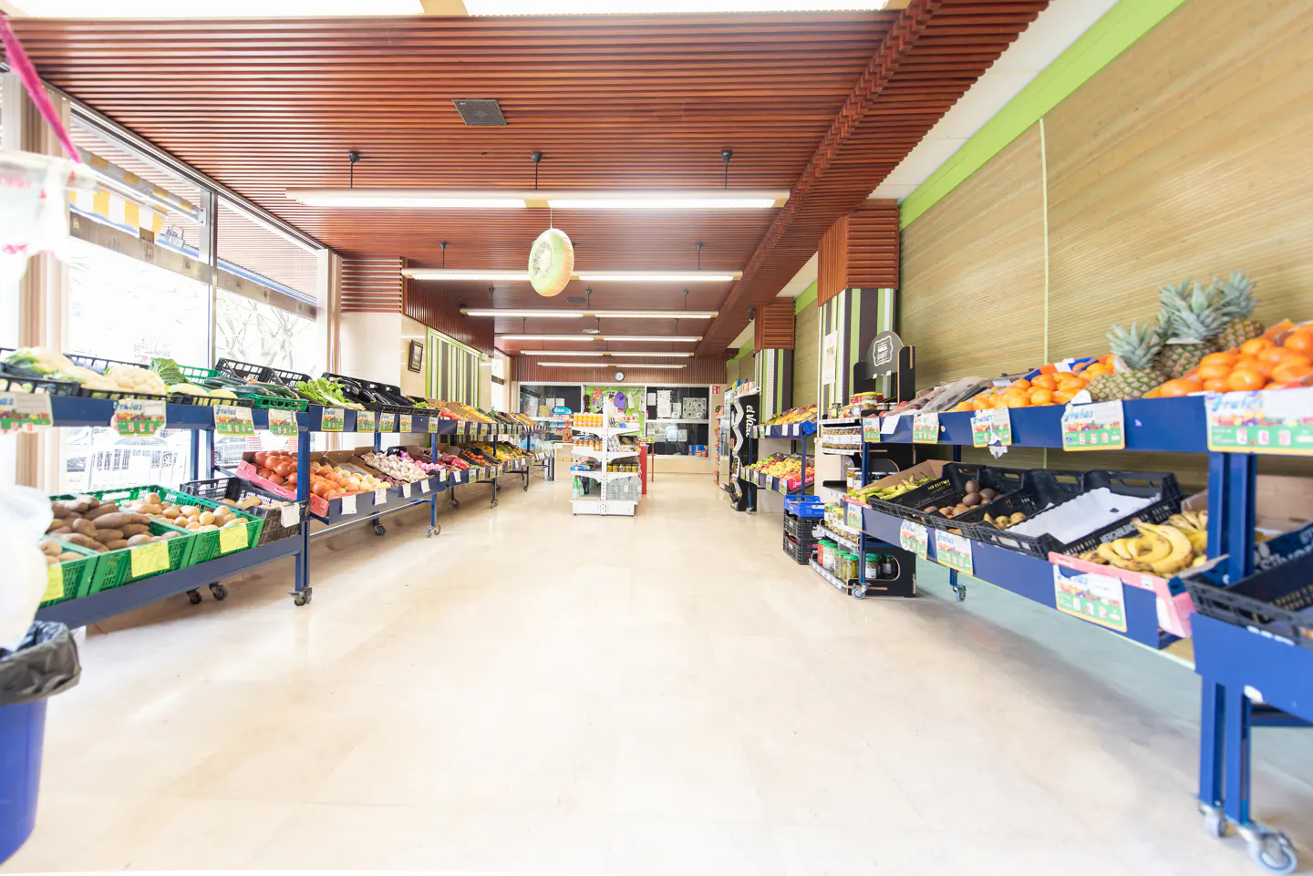 Interior view of a grocery store with produce on blue carts. The ceiling is wood with fluorescent lights.