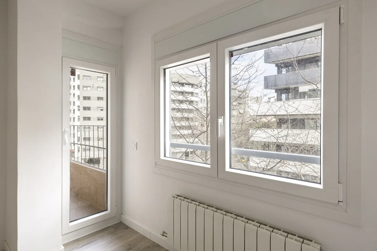 Bright, empty room with white walls, wood floor, radiator, and windows. Balcony door shows city view.