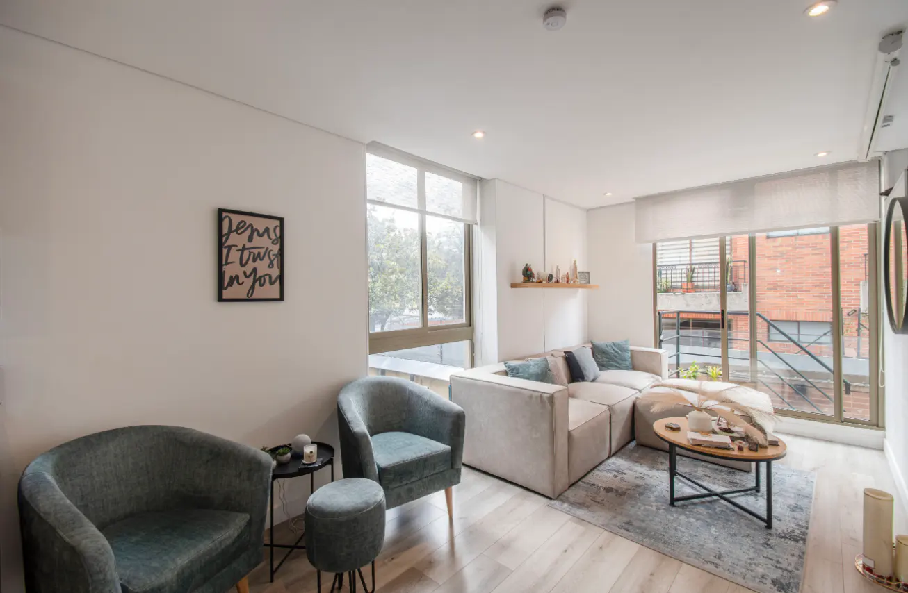 Bright living room with light wood floors, a beige sectional sofa, two blue armchairs, and large windows. A framed print hangs on the wall.