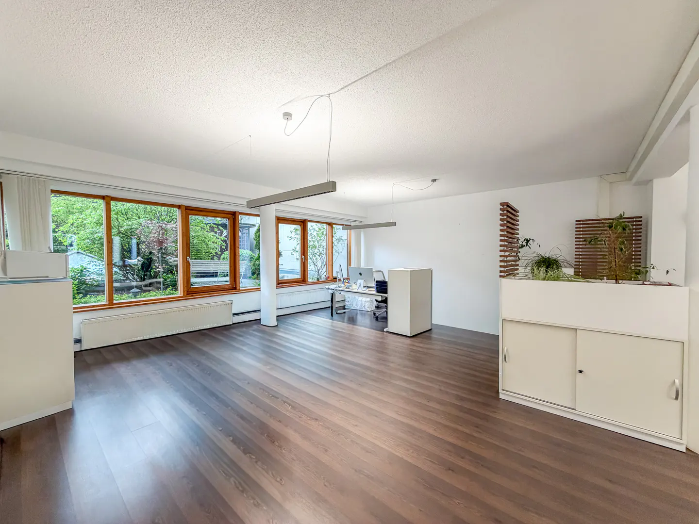 Bright, empty office space with wood floors, white walls, and large windows overlooking greenery. Desks and cabinets are also white.