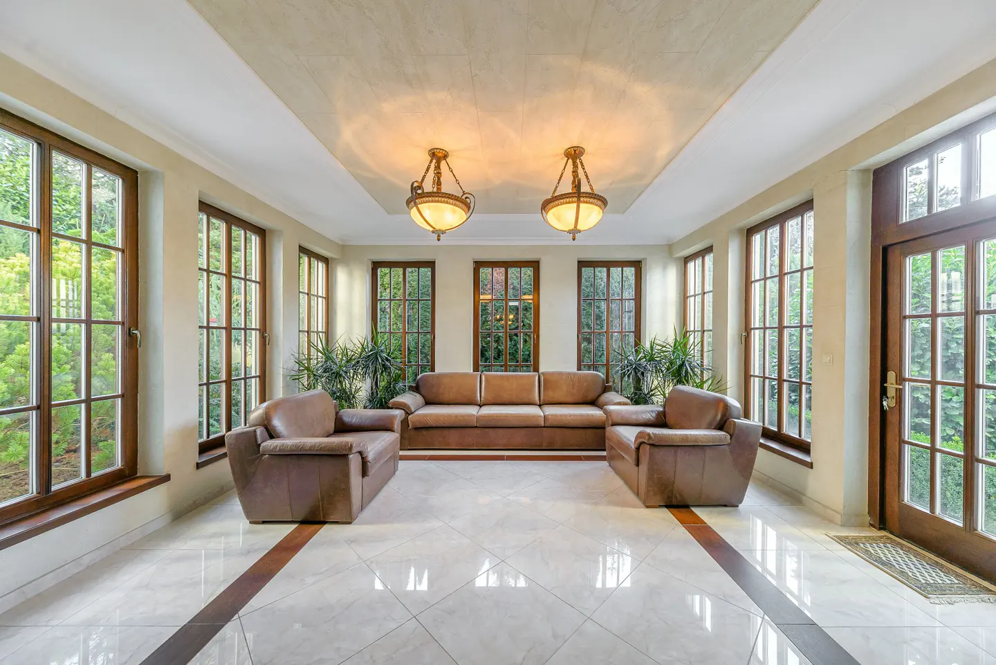 Bright sunroom with brown leather sofa and chairs, marble floors, and large windows overlooking greenery. Two ornate chandeliers hang from the ceiling.