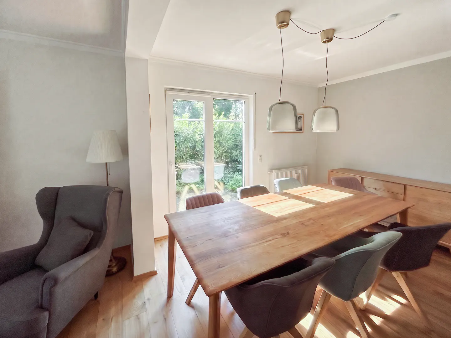 A bright dining room with a long wooden table and chairs, lit by pendant lights. A gray armchair sits near a window overlooking a green garden.