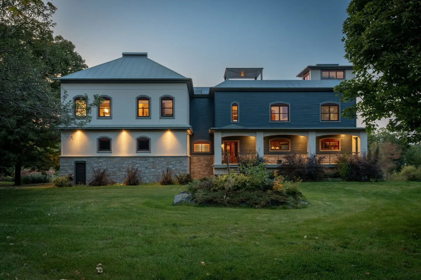 Two-story house with a white and blue exterior, set on a green lawn at dusk. Windows glow with warm light.