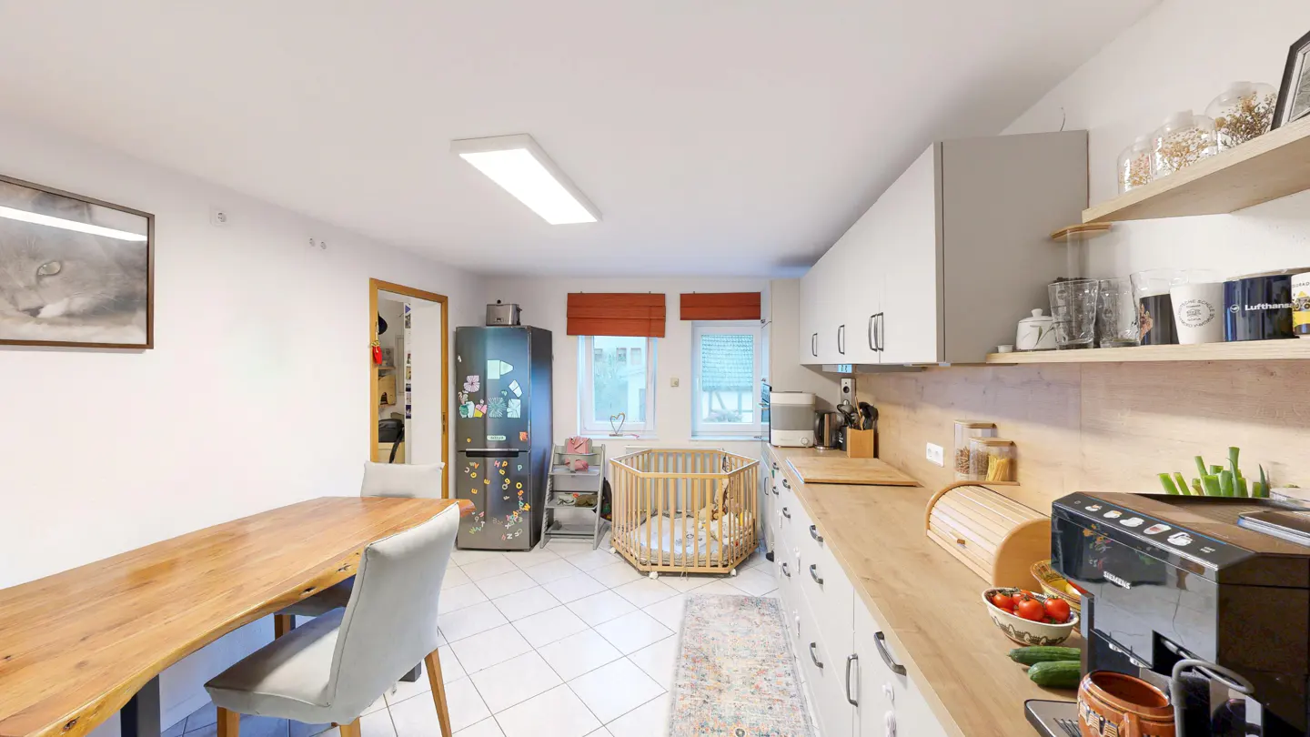 Bright kitchen with white cabinets, wood countertops, and a wooden dining table. A baby playpen sits near the window.