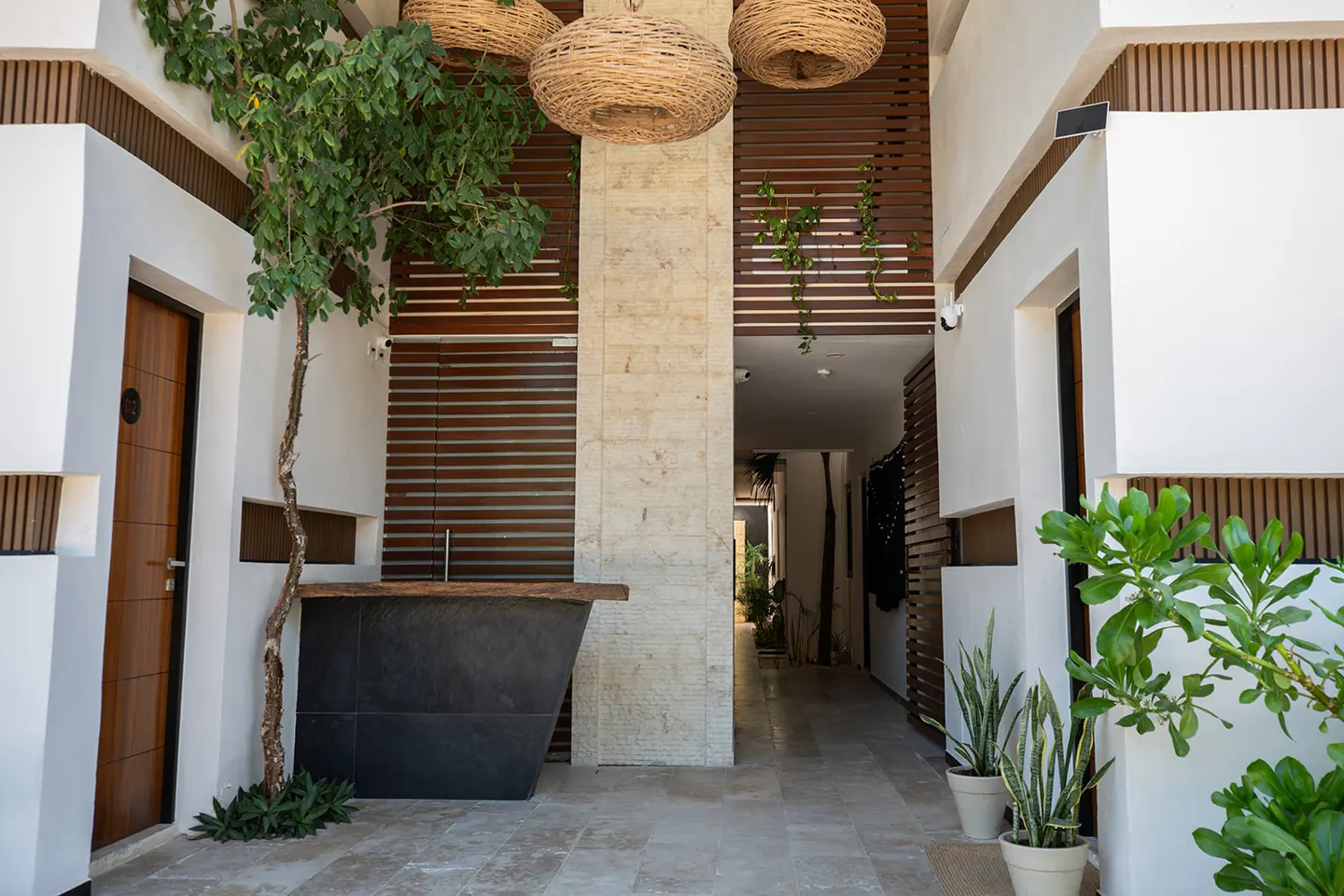 Hotel lobby with white walls, brown wood accents, and woven pendant lights. A black reception desk sits near a small tree.