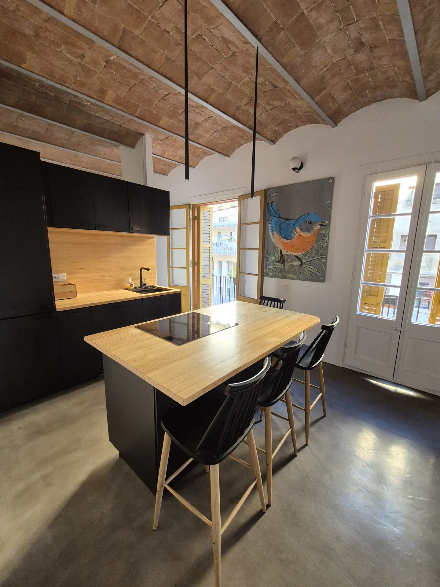 A modern kitchen with black cabinets, a light wood island with black chairs, and a brick ceiling. A blue bird painting hangs on the wall.
