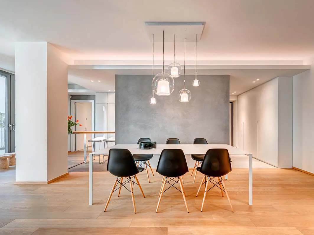 Bright dining room with a white table, black chairs, and glass pendant lights. Wood floors and a gray accent wall add warmth.