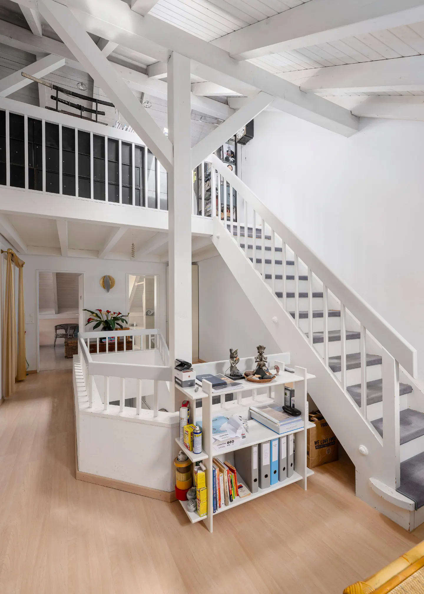 Bright, airy interior with white wooden beams, stairs, and shelving. Light wood floors and a glimpse into other rooms create an open feel.