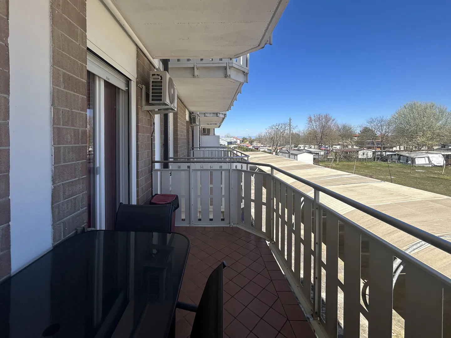 Balcony view with a black table and chairs on a tiled floor, overlooking a park with trailers under a blue sky.