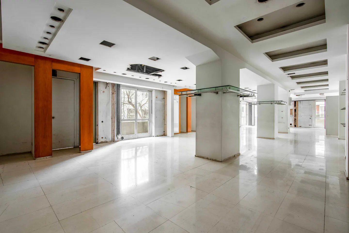 Empty commercial space with white marble floors, white walls, and orange accents. Columns with glass shelves line the space.