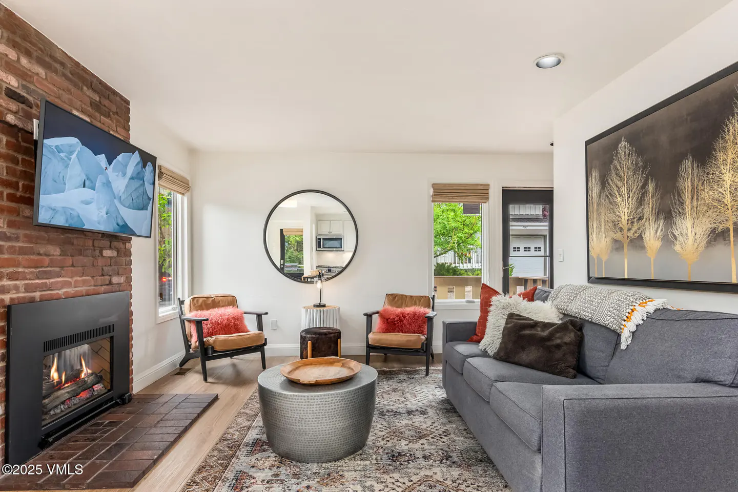 Living room with a gray sofa, two chairs, and a fireplace with a TV above it. A round mirror reflects the kitchen.