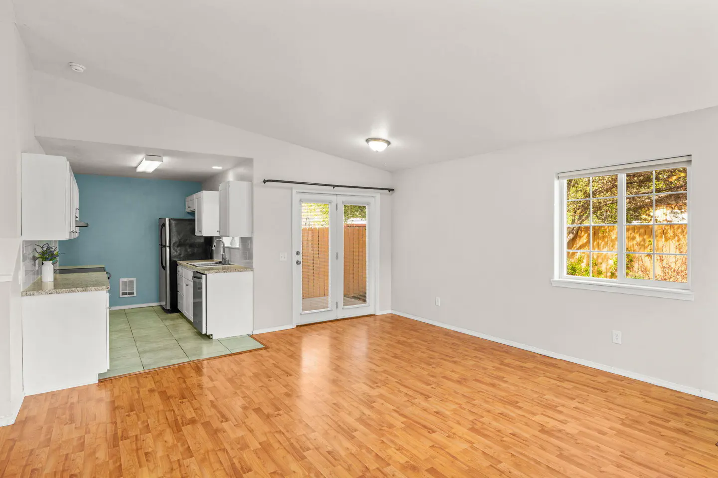 Bright, open-concept living space with wood floors, white walls, and a kitchen with blue accent wall. Patio doors and a window provide natural light.