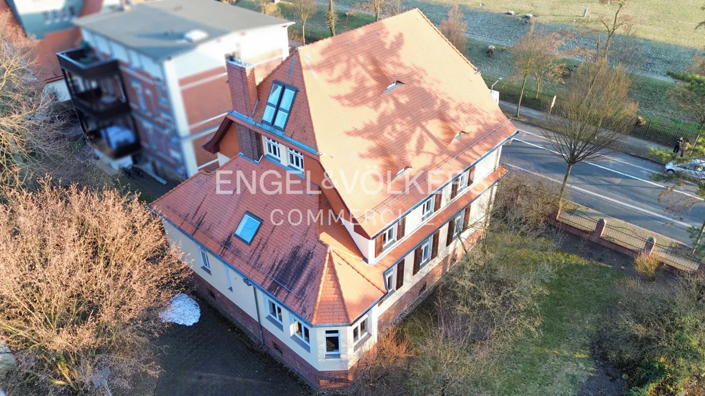 Aerial view of a three-story building with a red tile roof and white trim, surrounded by trees and a street.