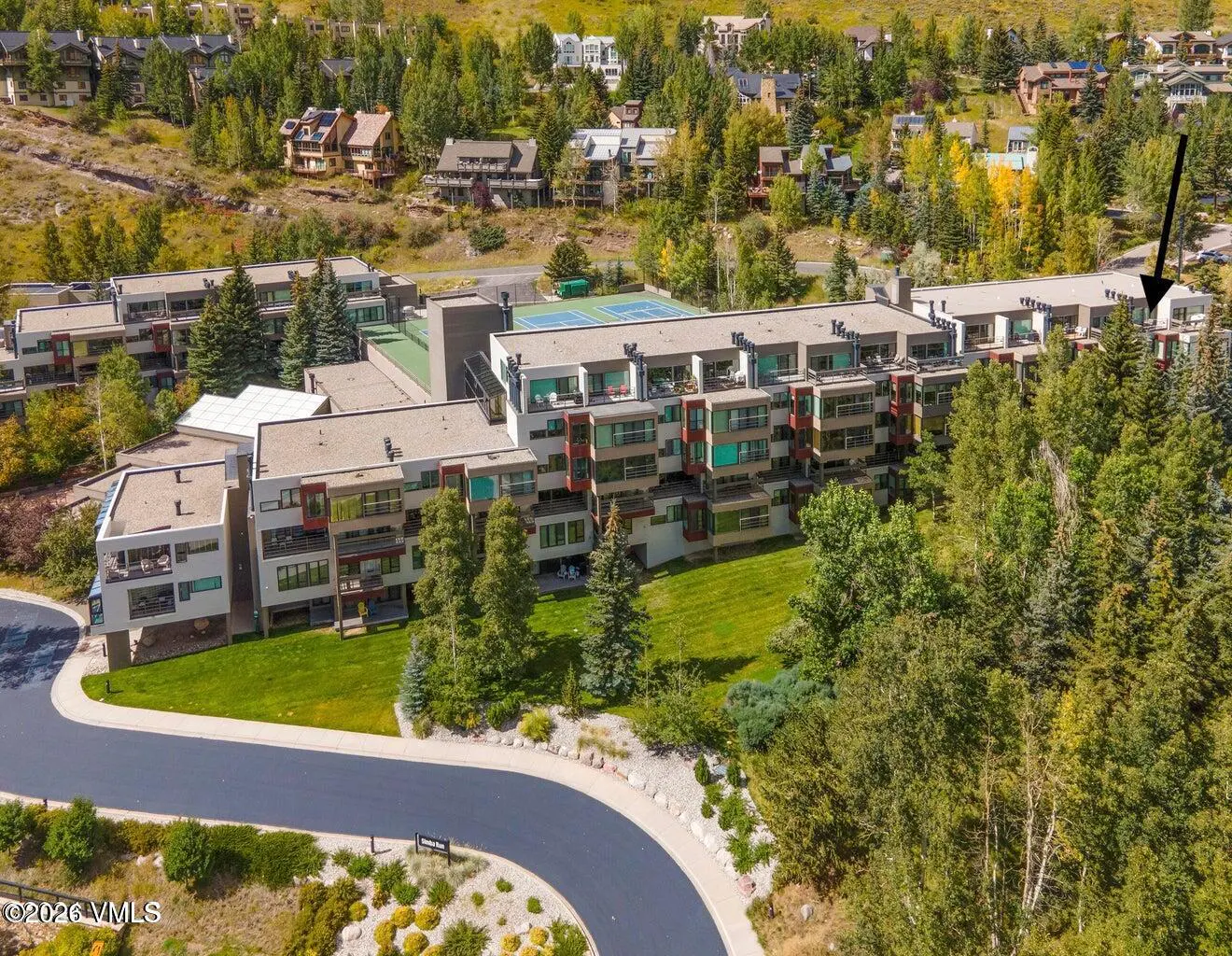 Aerial view of a multi-story condo building with a tennis court on the roof, surrounded by green trees and grass.