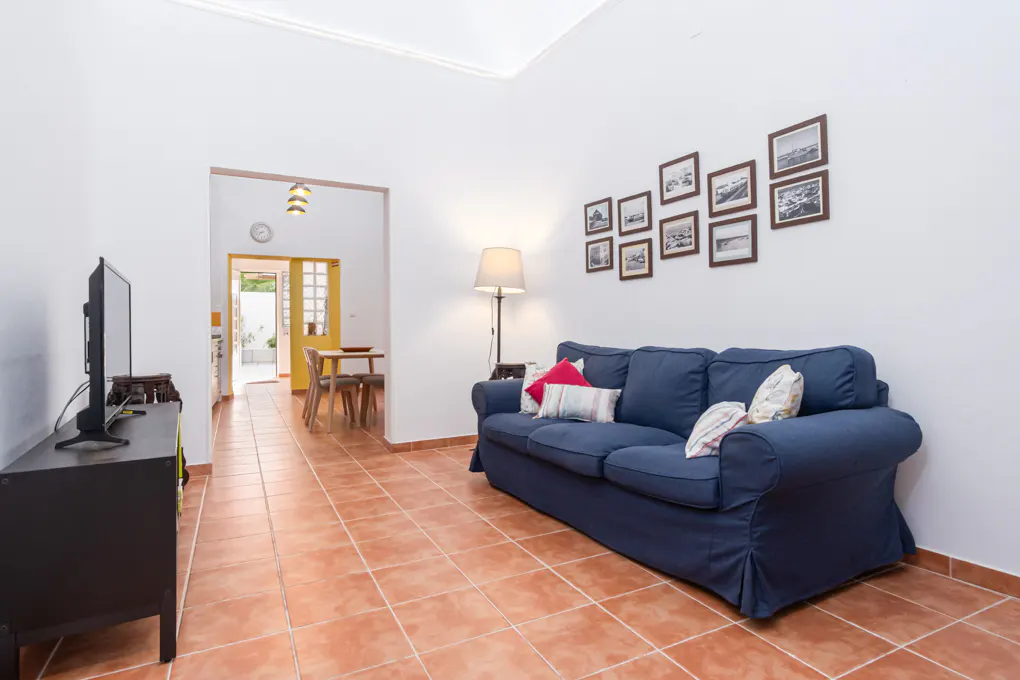 Living room with terracotta tile floor, blue sofa with pillows, and framed pictures on a white wall. A TV stands on a black cabinet.