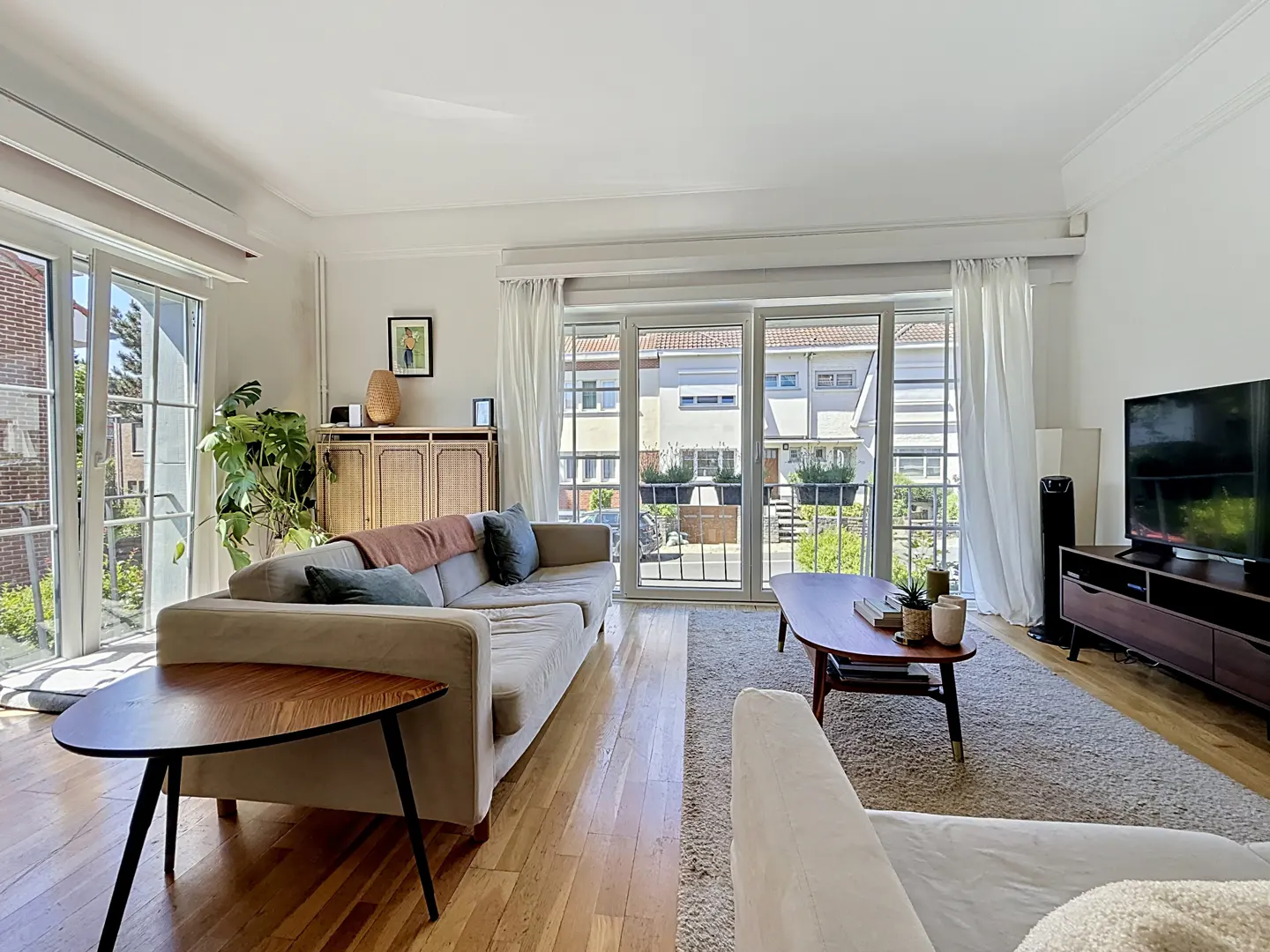 Bright living room with a beige sofa, wood floors, and large windows. A wooden cabinet and TV stand add warmth to the space.