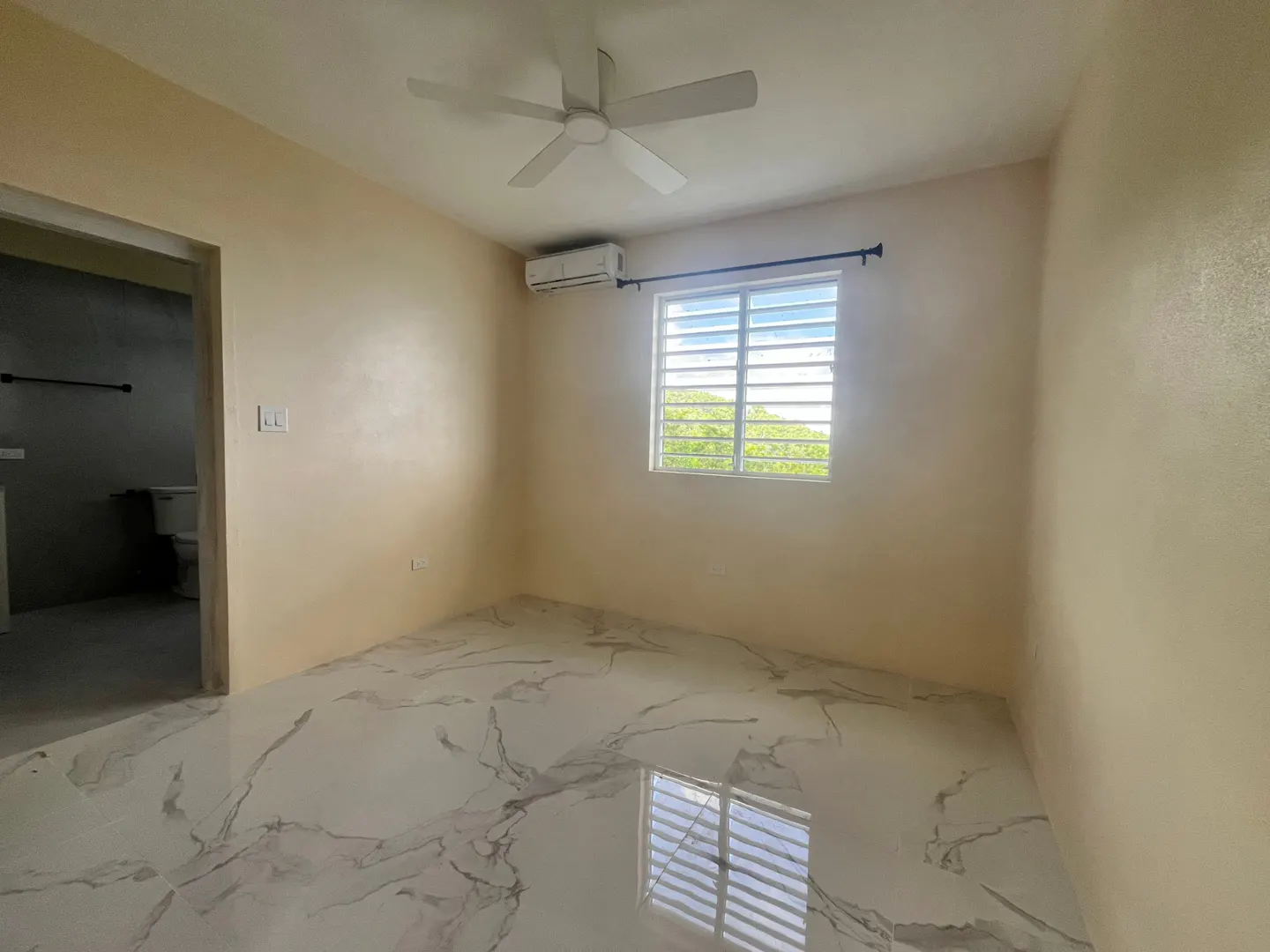 A bedroom with beige walls, a white ceiling fan, and marble floors. A window with white blinds is on the back wall. A bathroom is visible through a doorway.