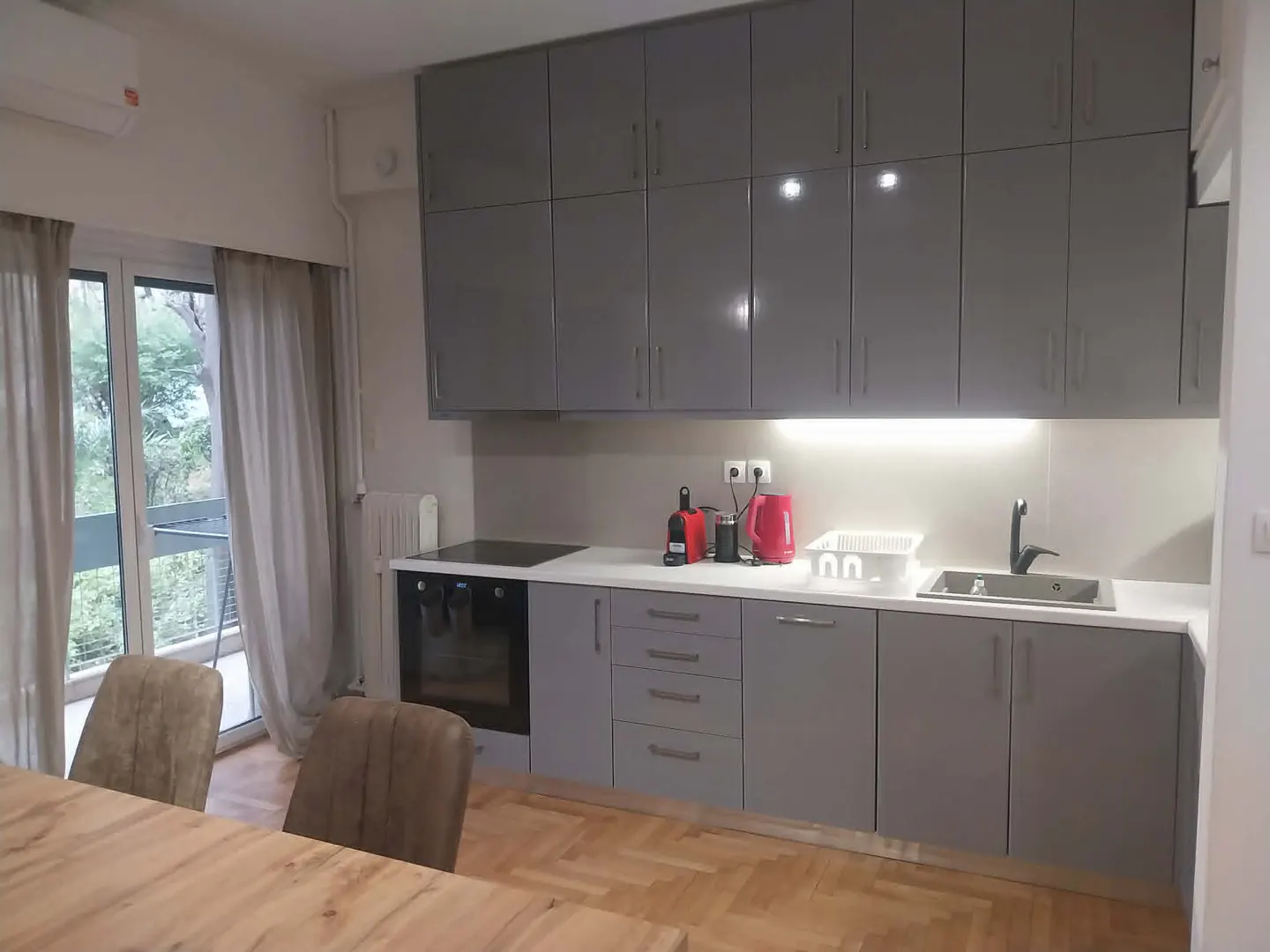 Modern kitchen with gray cabinets, white countertops, and a wooden table with chairs. A window with curtains overlooks greenery.